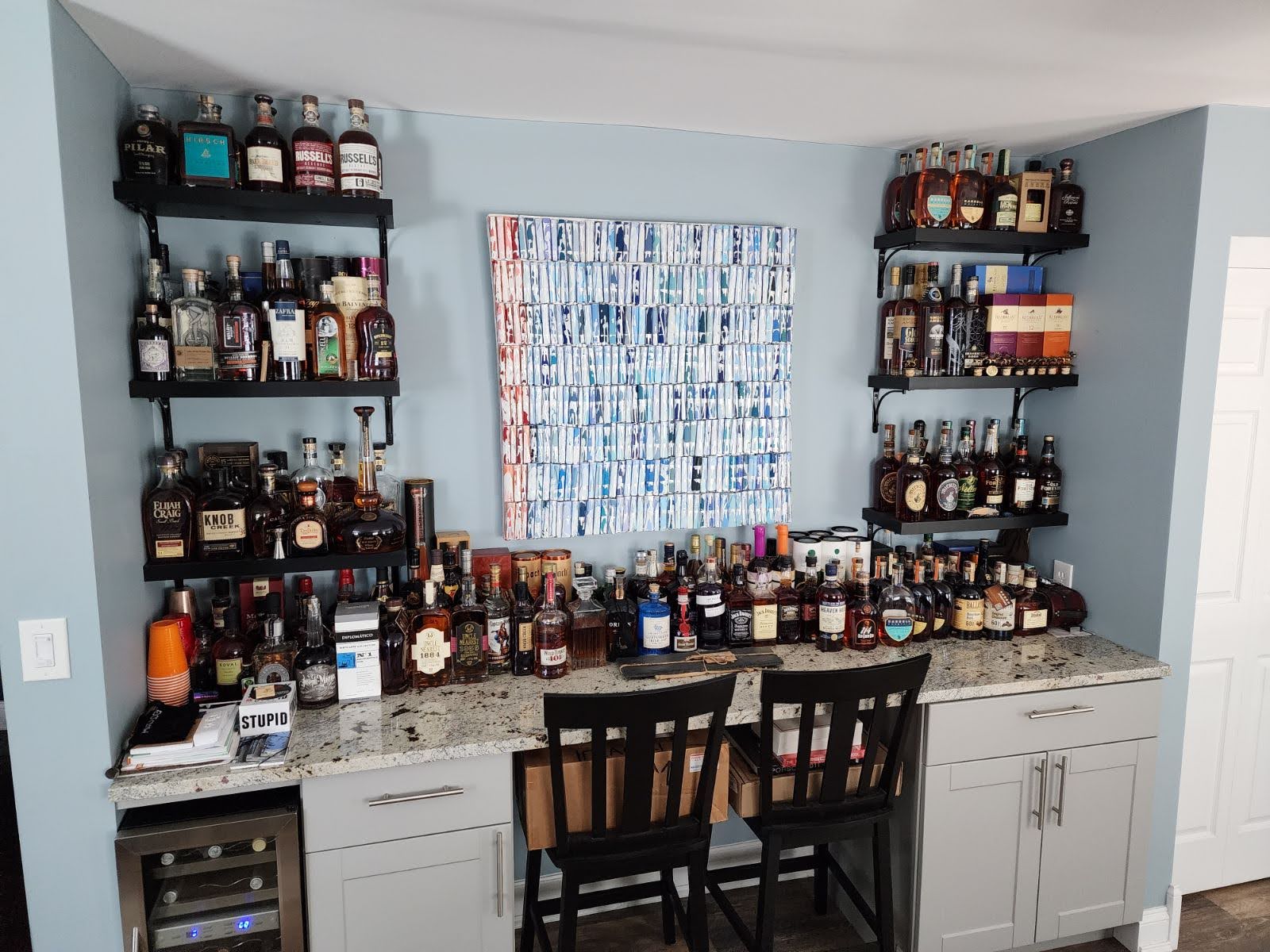 A home bar wall of whiskey bottles on black shelves above a granite counter — the collection that inspired the name.